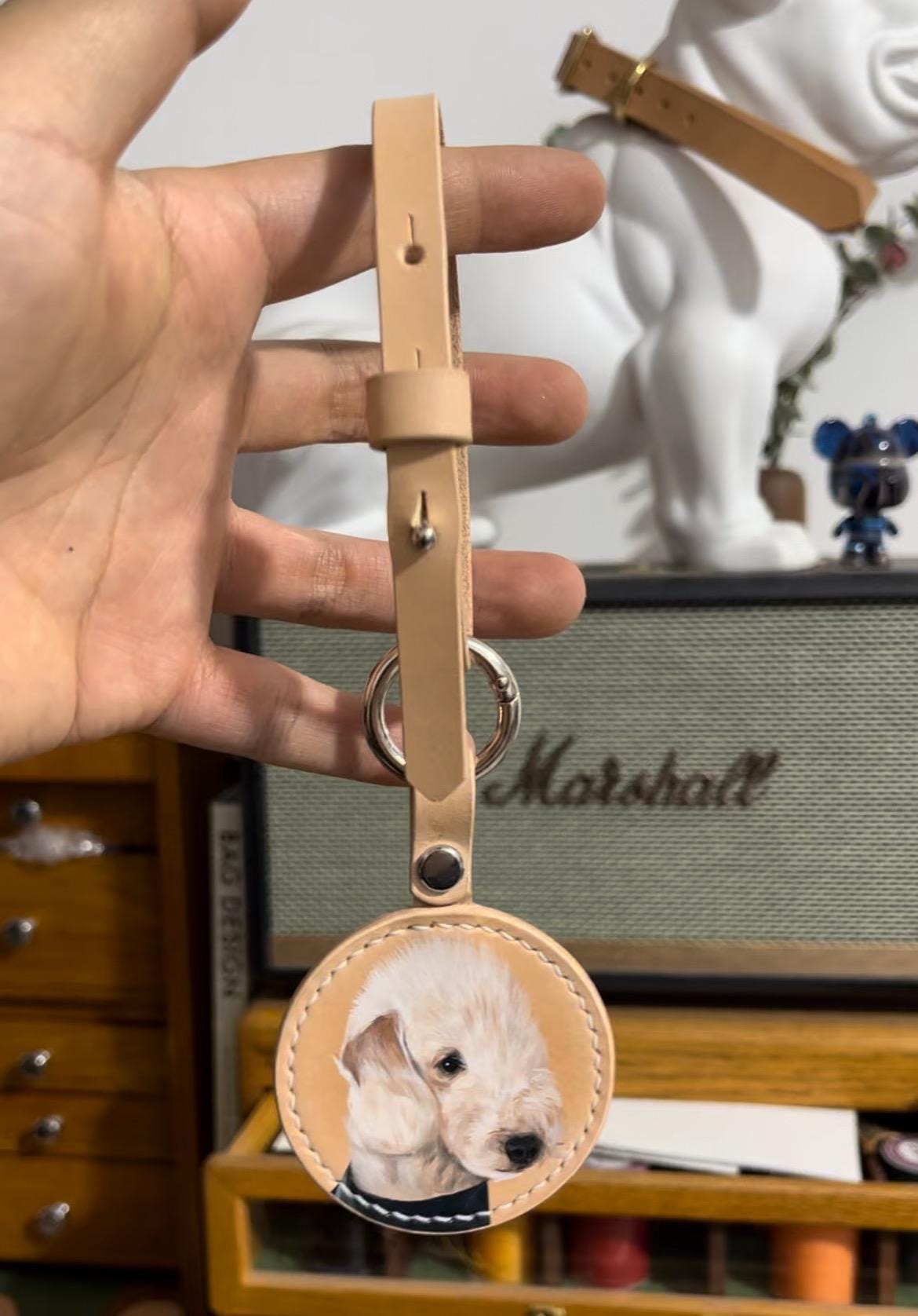 A handcrafted leather luggage tag featuring a custom hand-painted portrait of a white puppy, displayed in a workshop setting.