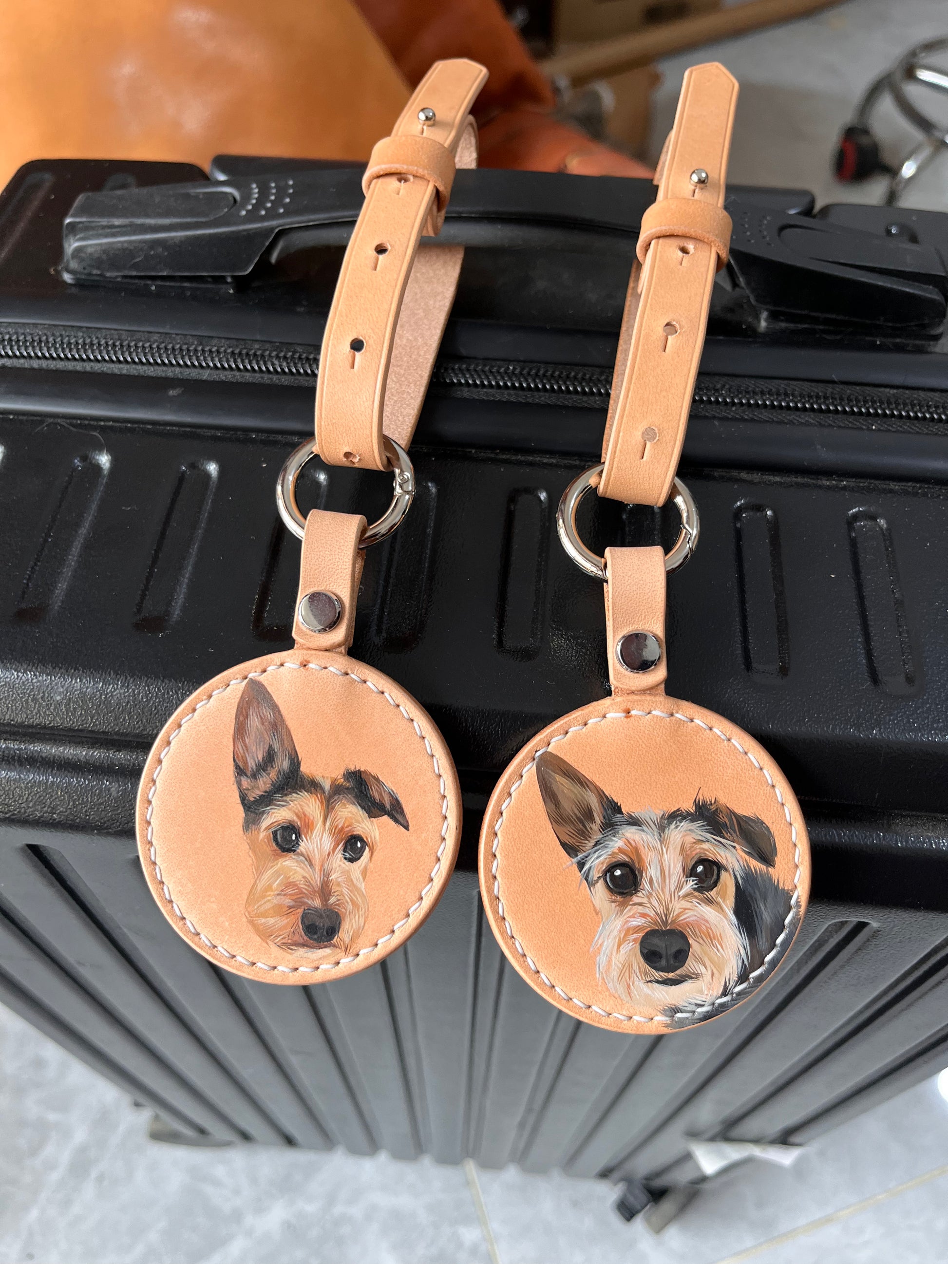 A pair of personalized leather luggage tags with hand-painted dog portraits, attached to a black suitcase, highlighting the unique customization.