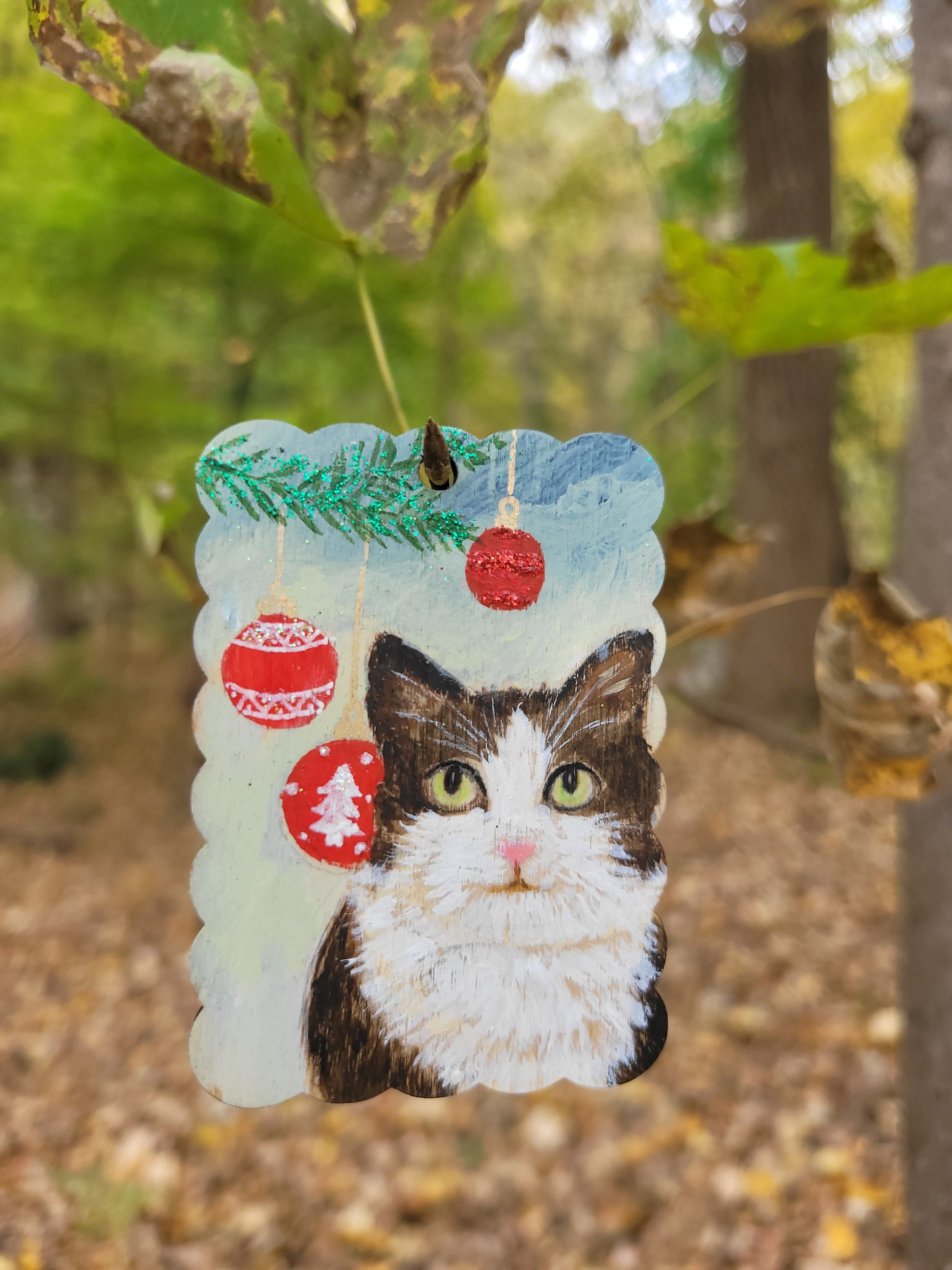Hand-painted cat portrait Christmas ornament wood tag hanging on a branch, featuring red ornaments.