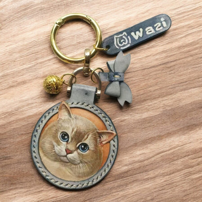 A leather keychain with a painted cat face, stitched border, gold ring, bell, blue bow, and a tag engraved "wazi" on a wooden background.