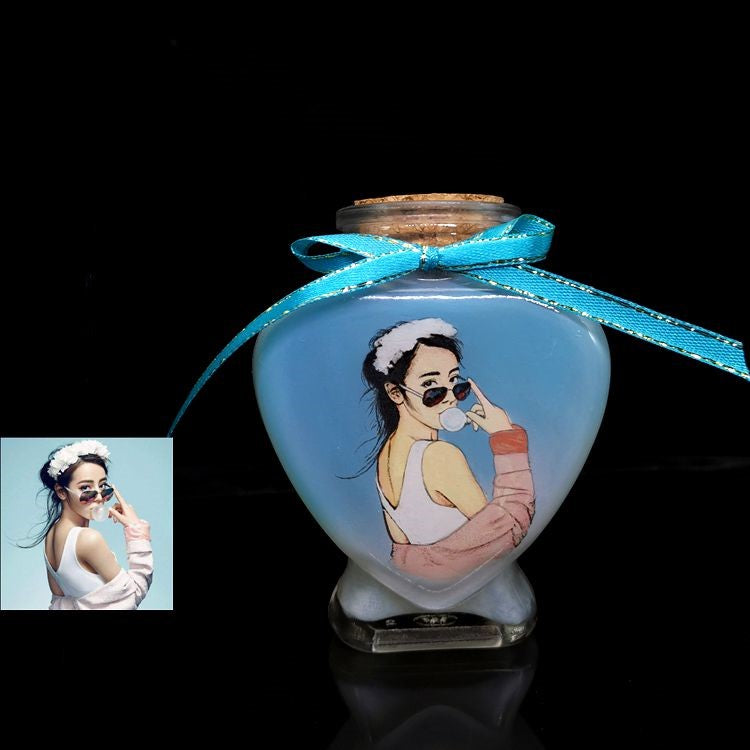 A heart-shaped hand-painted sand bottle featuring a portrait of a woman with sunglasses and a headband, tied with a blue ribbon.