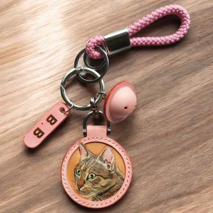 A pink leather keychain with a painted cat face, braided loop, metal keyring, and a tag with the letter “B” on a wooden background.